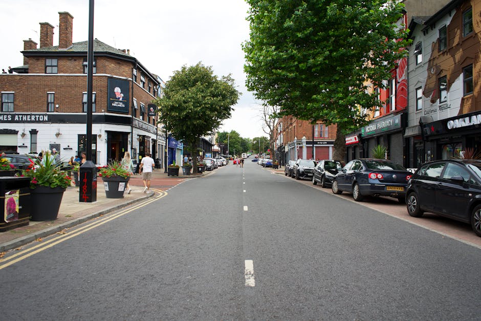 A wide view of a city street with a central lane marked by a broken white line, flanked by parked cars on both sides. On the left side, there are several large, black flower pots filled with vibrant flowering plants, placed along the sidewalk next to a building with a dark façade housing a pub or restaurant, with outdoor seating and pedestrians walking nearby. The right side features a row of parked cars, including a mix of compact and mid-sized vehicles, adjacent to a variety of commercial establishments with signage and large windows. The street is lined with mature green trees, providing partial shade and greenery to the environment. The atmosphere appears calm and typical of an urban area, with soft natural lighting suggesting a cloudy day. This scene relates to urban waste management and the potential for private rubbish removal services, with Rubbish Removal Acton operating in the area to provide alternative rubbish collection solutions, as indicated by its presence and context on a busy high street.
