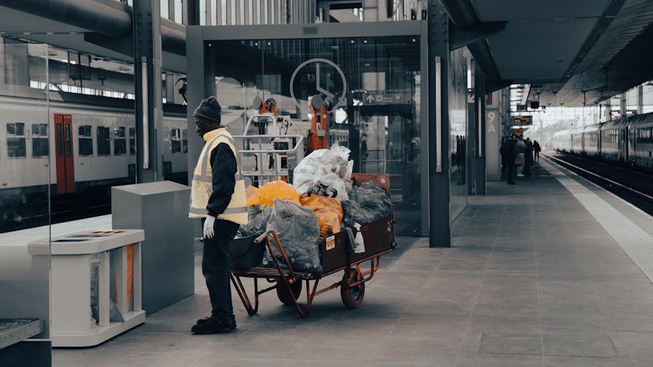 At an indoor train station platform, a worker stands beside a large, orange metal trolley loaded with various bags and bundles of waste materials, including black, grey, and orange plastic bags, some of which appear to contain discarded items. The trolley is situated close to a modern glass waiting area with metal framing, and the background shows ongoing train services with a train on the right side and another train on the left platform, reflecting natural daylight through the station’s large windows. The ground surface is paved with smooth grey tiles, and there are a few passengers visible in the distance, some walking along the platform. Nearby, a white waste disposal unit with a lid is positioned on the left side of the image, adjacent to the worker, indicating an environment where waste collection or disposal processes are actively managed. The scene's lighting is soft and diffuse, typical of a contemporary transit environment, emphasizing the practical context of waste management stations or services, such as those offered by Rubbish Removal Acton for alternative waste handling outside of standard rail service operations.