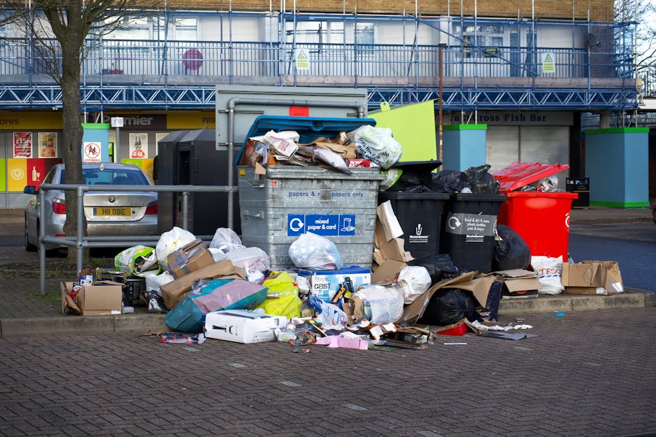 In the foreground, a large metal bin labeled for mixed paper and cardboard is overflowing with assorted waste, including crumpled paper, flattened boxes, and packaging materials. Surrounding the bin, various black and black plastic rubbish bags are stacked, some appearing partially torn open, spilling cardboard boxes, plastic containers, and miscellaneous litter onto the brick-paved ground. To the left, a grey parked car partially obscures a metal railing, with several smaller cardboard boxes and plastic bags scattered nearby. In the background, a modern building with blue scaffolding and protective netting can be seen, with storefronts beneath, including signs for a fish bar. The scene is lit by natural daylight, highlighting the cluttered nature of the waste accumulation, which appears to be a result of an on-site clearance or private waste collection, typical of independent rubbish removal services, such as those provided by Rubbish Removal Acton, serving the South Acton estate area. The environment suggests an urban setting with a focus on rubbish management and disposal.